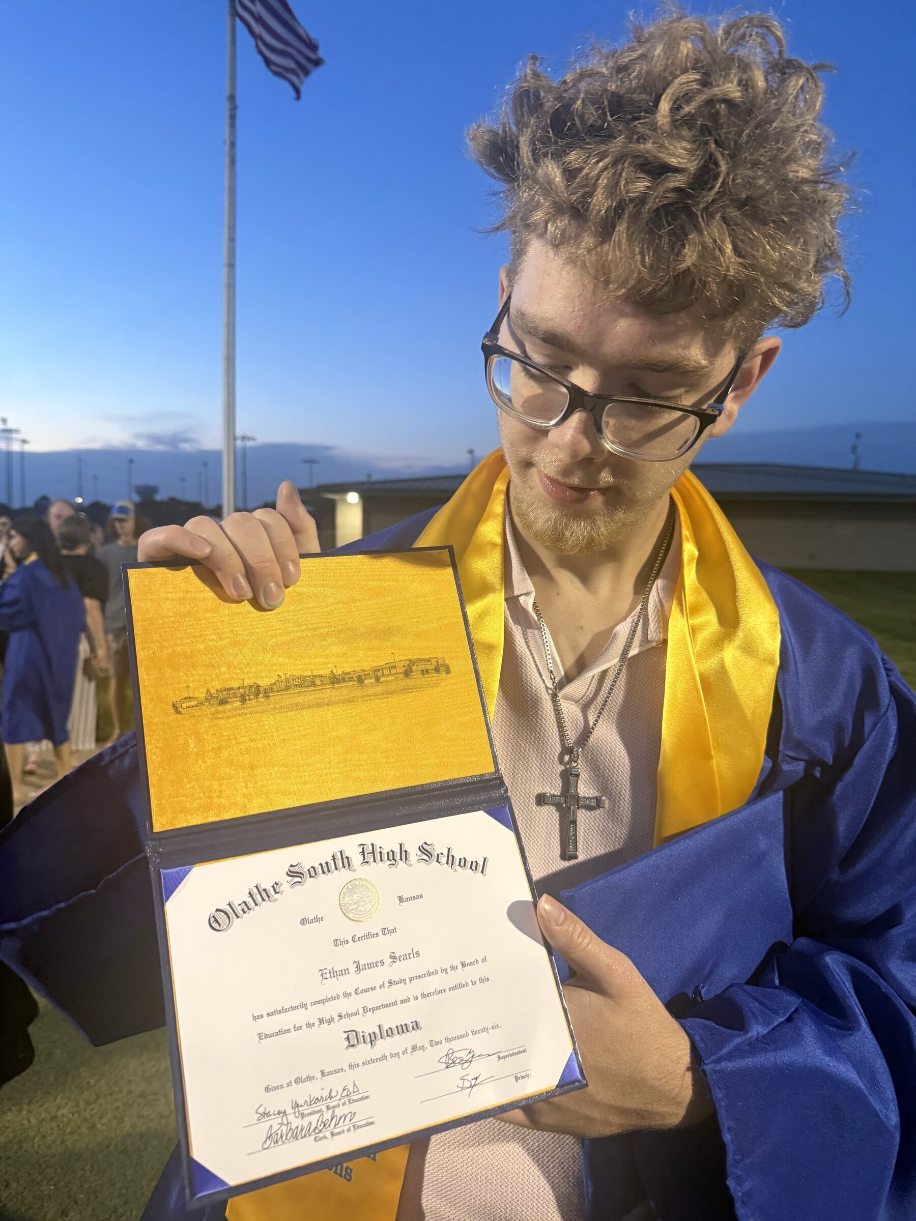 Ethan in his graduation gown holds his open diploma at dusk, a cross necklace catching the light.