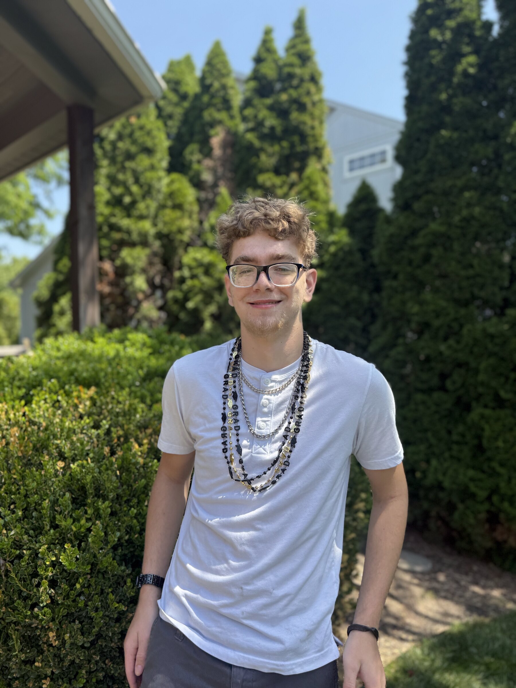 Ethan stands outdoors in a white t-shirt and layered necklaces, looking calmly into the camera.
