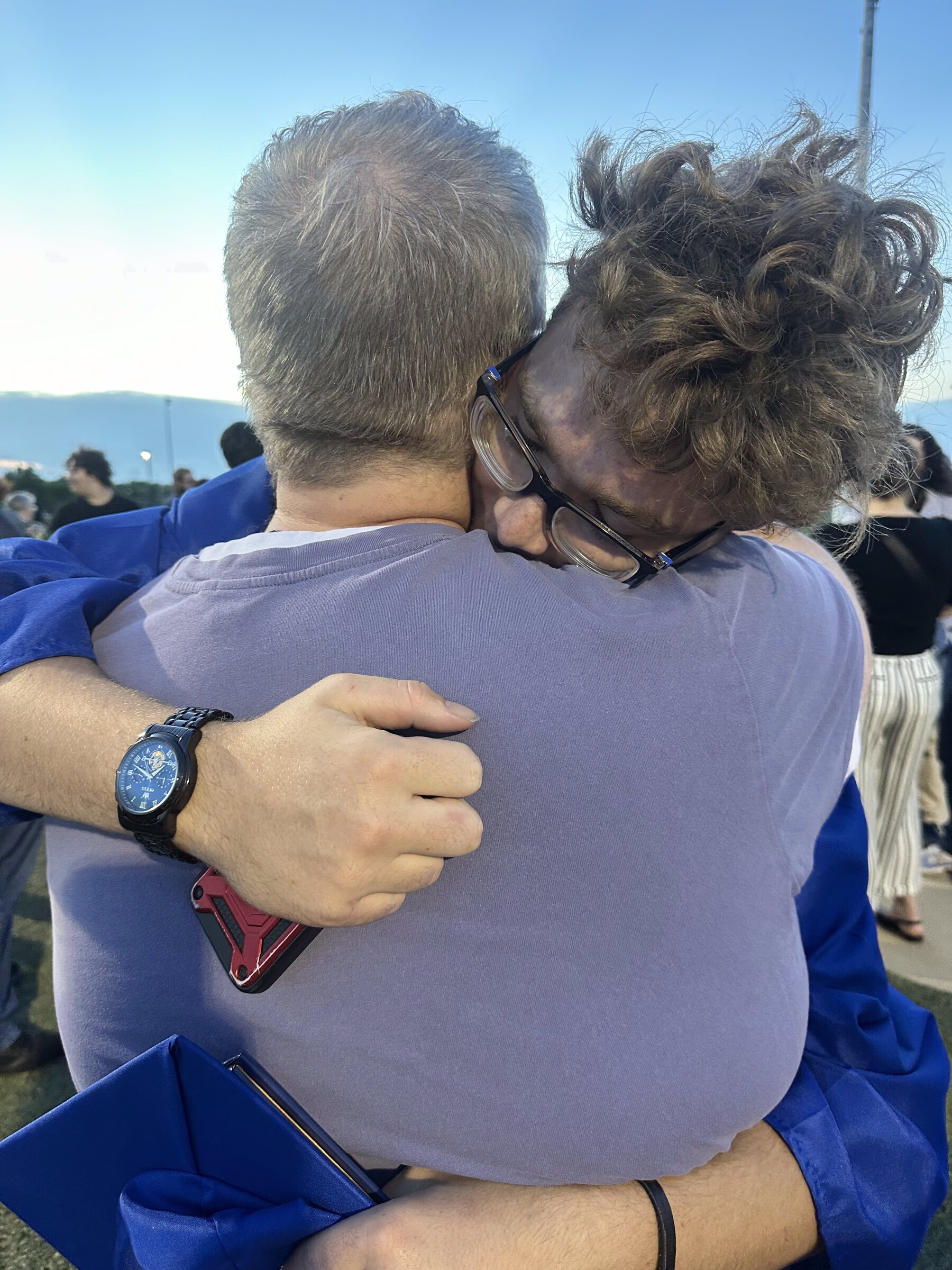 Jacob holds Ethan in a long embrace at his graduation. Ethan's curly hair against his father's shoulder; a watch on Jacob's wrist.