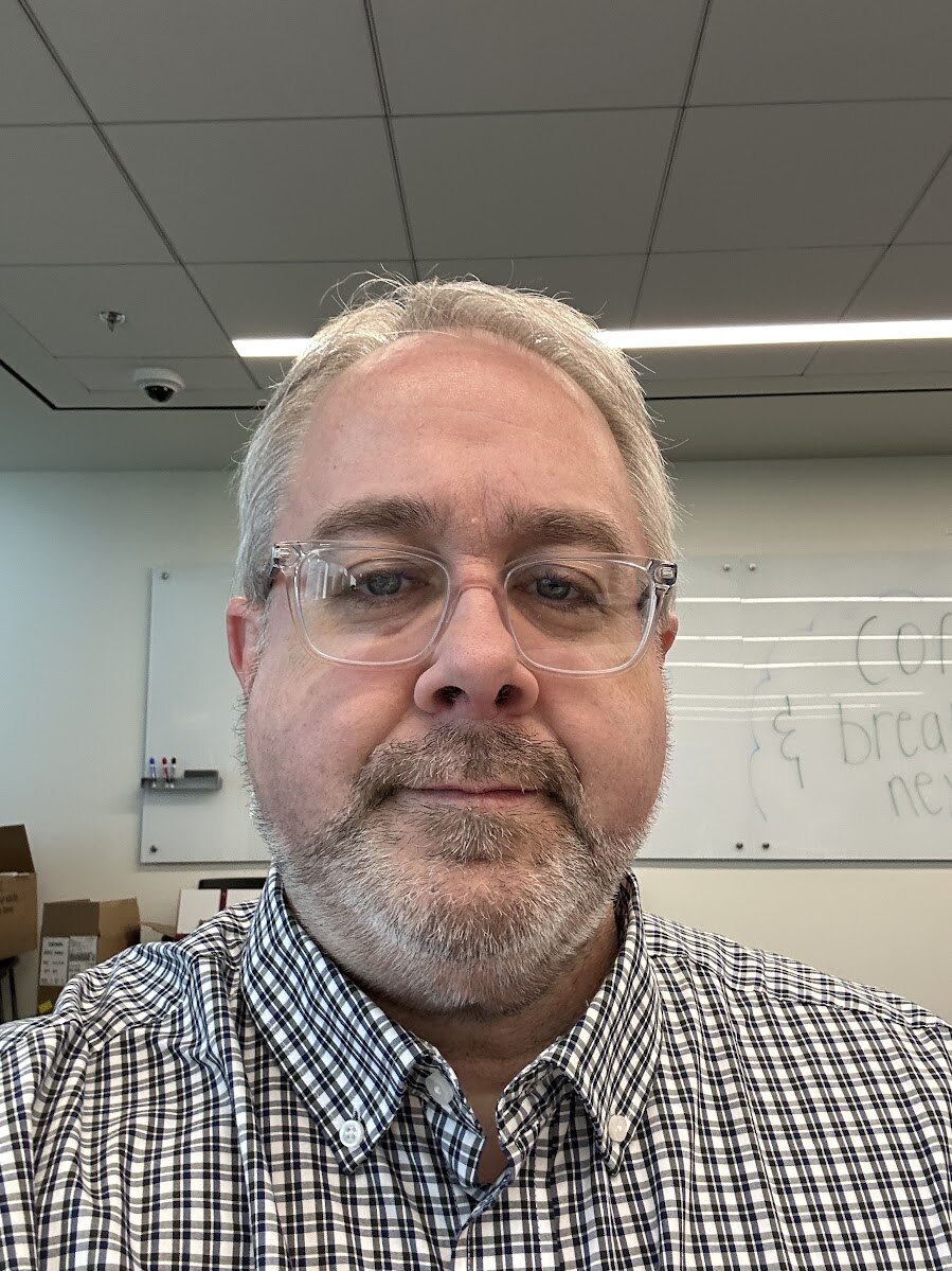 Jacob, mid-forties, in clear-frame glasses and a checkered button-down, standing in front of a whiteboard at work.